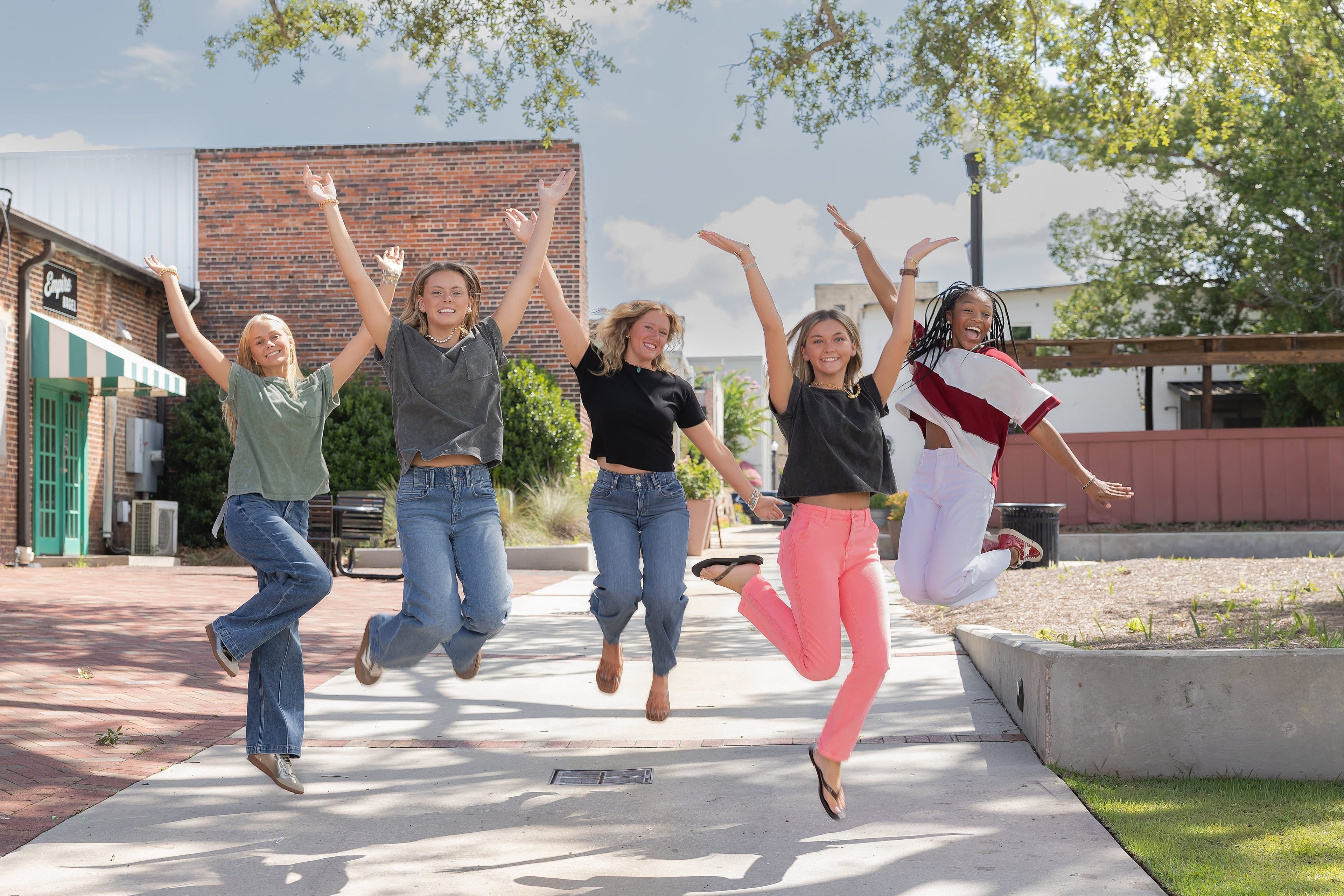 Five young women jump joyfully on a sunny outdoor sidewalk in front of brick buildings and trees. They are wearing casual everyday clothes, with one person wearing bright pink pants and another in a white top with a red stripe.